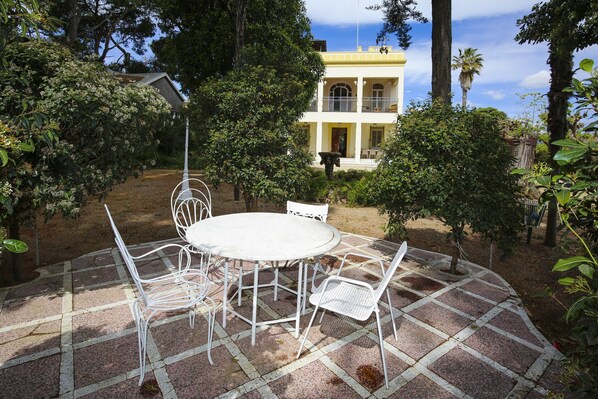 Un patio con una mesa redonda y cuatro sillas, rodeado de un jardín con diversas plantas y árboles.