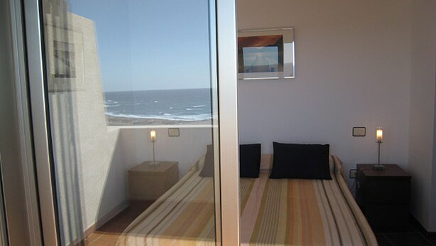 A hotel room with a large window overlooking the ocean, a bed with striped bedding, two bedside tables with lamps, and a small framed picture on the wall. - Modern Oceanfront Penthouse Apartment. Very Spacious. Big Sunbathing Terrace. (El Medano)