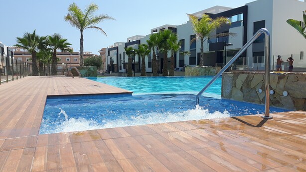 Zwembad met houten terras, palmbomen en moderne gebouwen op de achtergrond.
