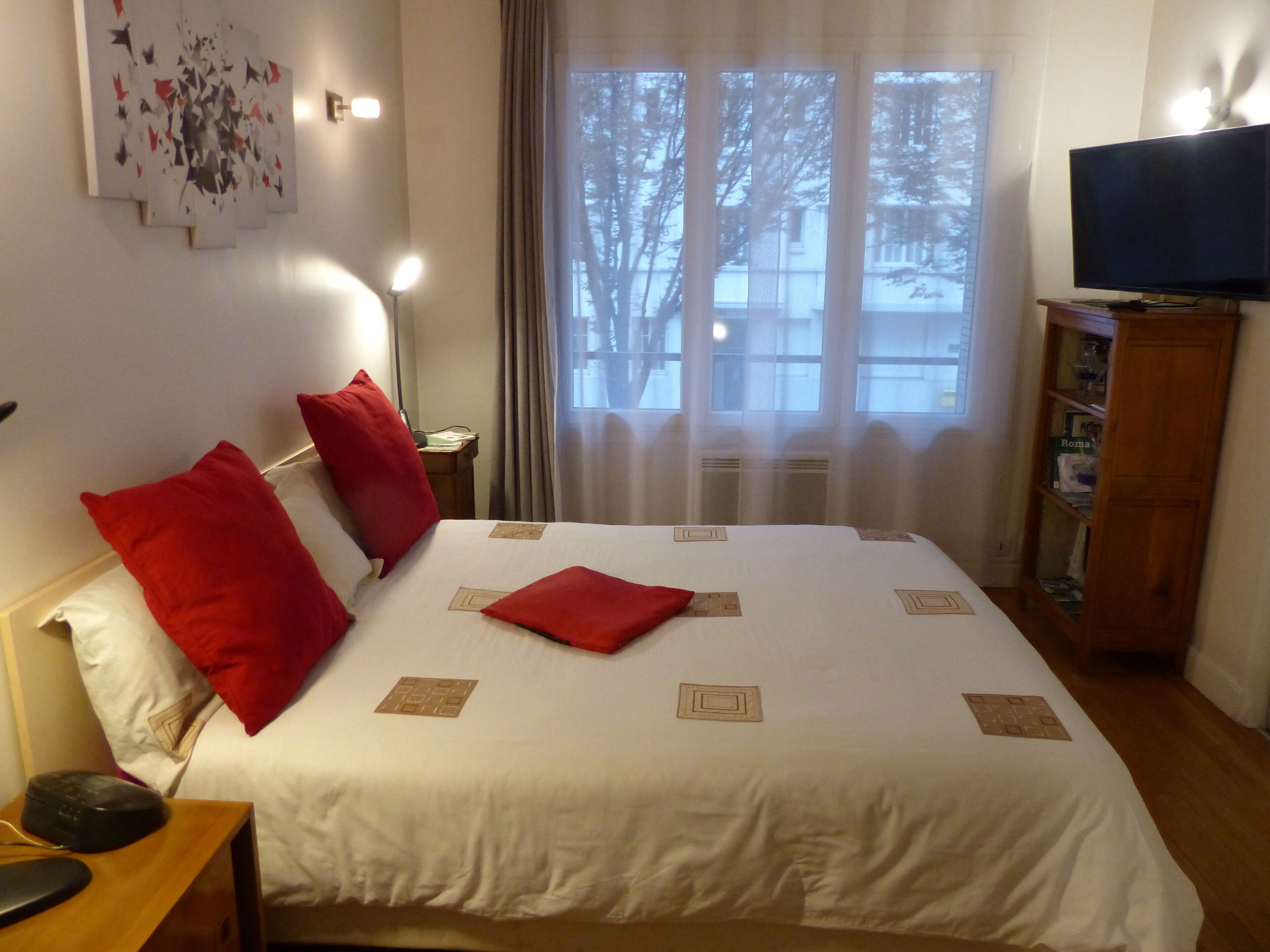Et hotelværelse med seng, røde puder, et fladskærms-tv, et natbord med lampe og en bogreol.