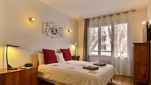 Une chambre à coucher comprenant un lit, deux tables de chevet, une commode, une armoire en bois et une fenêtre avec des rideaux.