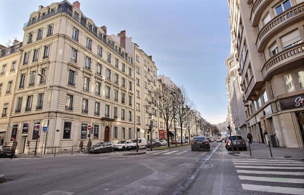 A street view with multi-story buildings, parked cars, and a pedestrian crossing.