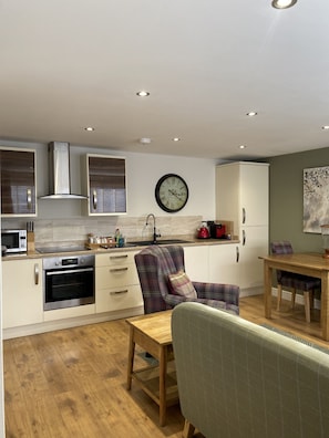 A modern kitchen with built-in appliances, a dining area with a wooden table and chairs, and a clock on the wall. - Brewers Cottage - sleeps 2 guests  in 1 bedroom (Foolow)