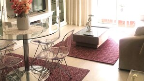Una sala de estar moderna con una mesa de comedor de vidrio, sillas transparentes, un televisor de pantalla plana, un mueble con puertas de vidrio y un ventanal con cortinas.
