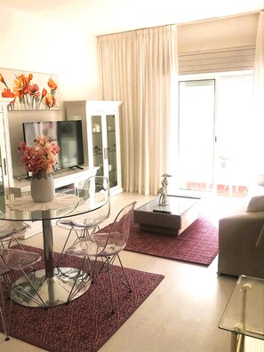A modern living room with a glass dining table, clear chairs, a flat-screen TV, a cabinet with glass doors, and a large window with curtains.