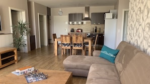 A modern living room with a wooden coffee table, a beige sofa with a teal pillow, and a dining area with wooden chairs and a kitchen in the background. - Comfortable holiday apartment within walking distance of the beach, Fehmarn Golf Park and surfing area  (Fehmarn)