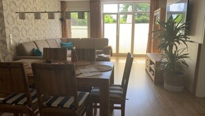 A dining area with a wooden table and chairs, a sofa, and a large window with curtains. - Comfortable holiday apartment within walking distance of the beach, Fehmarn Golf Park and surfing area  (Fehmarn)