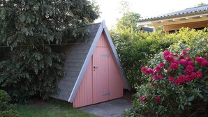 Een klein, roze tuinhuis met een schuin dak, omringd door groen en een struik met roze bloemen.