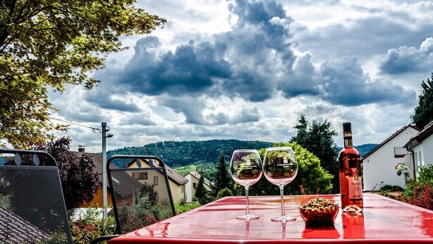 A table with two wine glasses and a bottle, set against a backdrop of a cloudy sky and a mountainous landscape.