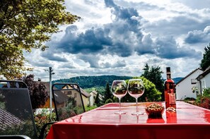 Ein Tisch mit zwei Weingläsern und einer Flasche, vor einer bewölkten Landschaft mit Bergen.