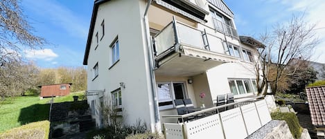 A two-story residential building with a balcony, a small garden, and a paved driveway.
