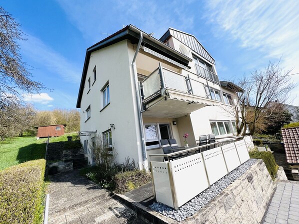 A two-story residential building with a balcony, a small garden, and a paved driveway.