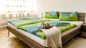 A neatly made bed with a green and blue striped bedspread, two pillows with green and blue patterns, and a tray with a teapot, cup, and pastries on the side.