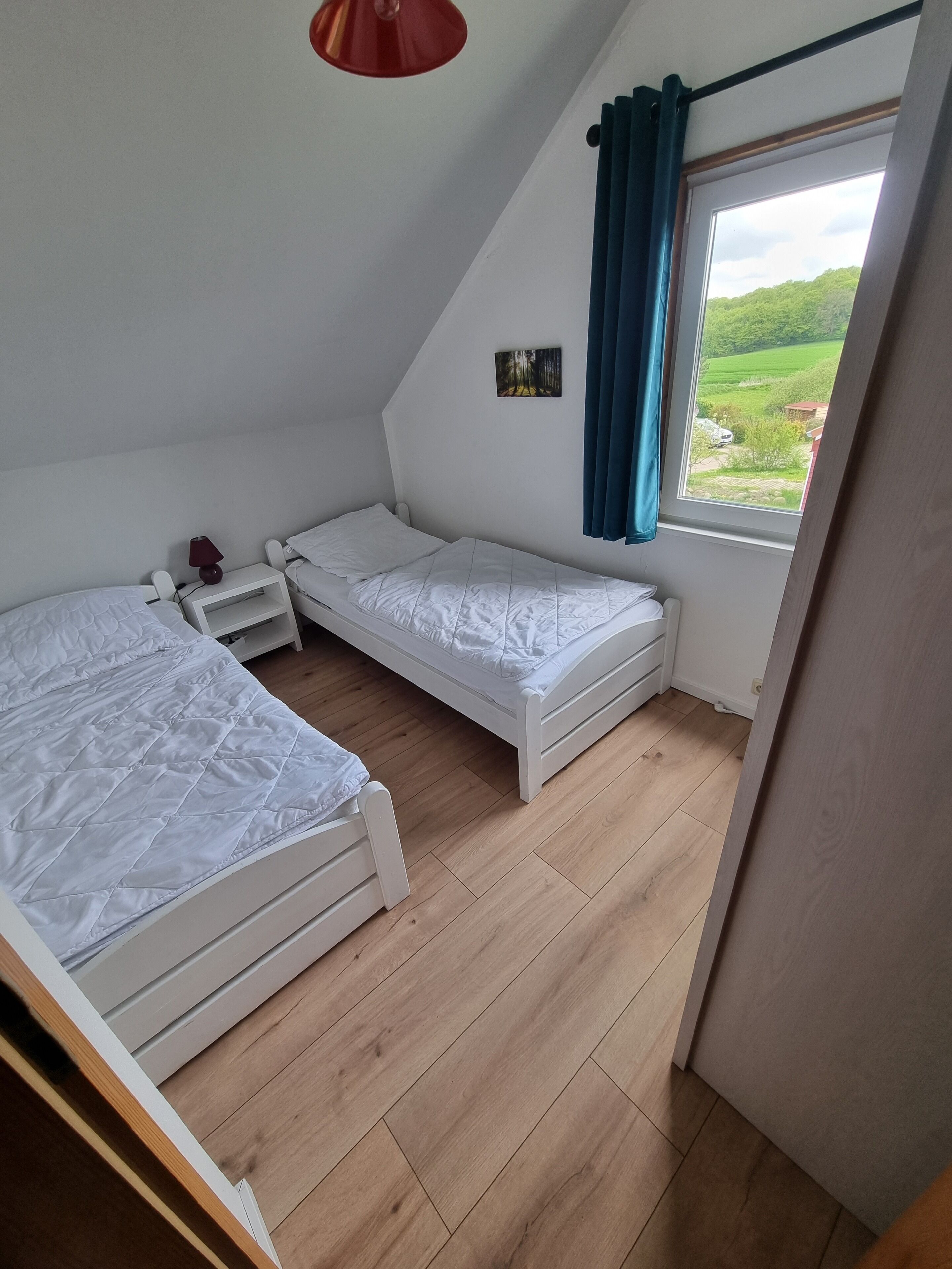 Una habitación pequeña y bien iluminada con dos camas individuales, una mesita de noche, una lámpara roja y una ventana con vistas a zonas verdes.