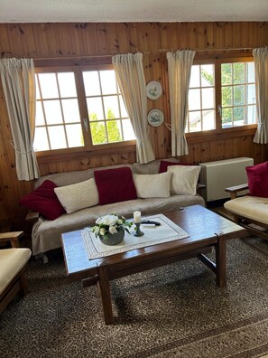 Una habitación acogedora con detalles de madera, un sofá con cojines rojos y blancos, una mesa de centro con una vela y flores, y vistas a través de amplios ventanales con cortinas.