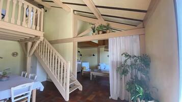 A room with a dining table, chairs, a staircase, a ceiling with wooden beams, and potted plants.