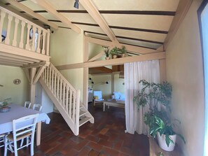 A room with a dining table, chairs, a staircase, a ceiling with wooden beams, and potted plants.