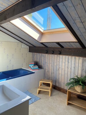 A room with a skylight, a bathtub, a small wooden stool, and a shelf with a plant.