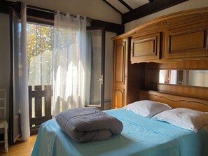 Een slaapkamer met een bed, houten hoofdbord en uitzicht op bomen door een raam met dunne gordijnen.