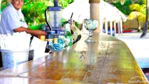 A bar with a wooden counter, a blender, and a glass. There are bottles and a bucket with straws. - Arenas Beach Hotel (Corn Islands)