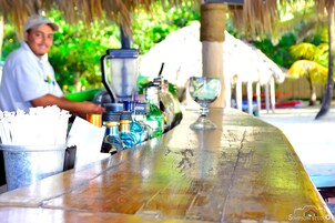 Un bar con mostrador de madera, una licuadora y un vaso. Hay botellas y un cubo con pajitas.