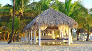 A beach hut with a thatched roof, surrounded by palm trees and white benches. - Arenas Beach Hotel (Corn Islands)