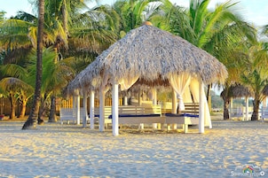 Una cabaña en la playa con techo de paja, rodeada de palmeras y bancos blancos.