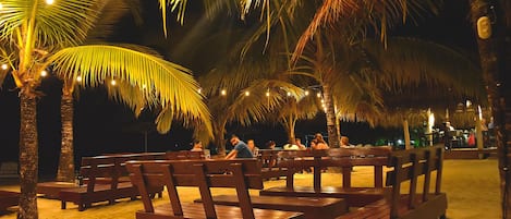 Un espace aménagé au bord de la plage, avec des bancs en bois et des palmiers, illuminé par des guirlandes lumineuses.