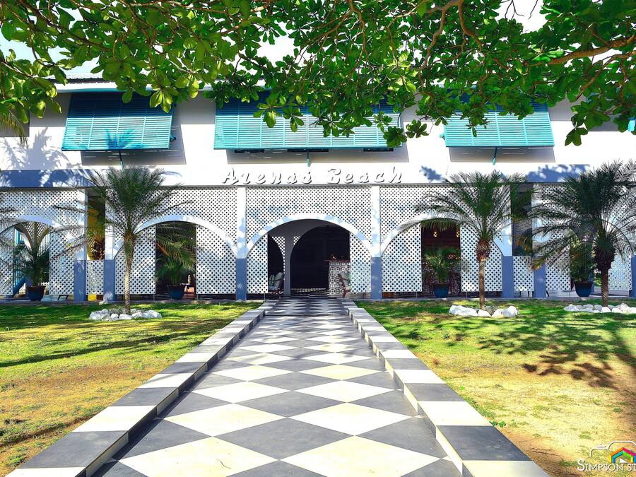 Entrada de un hotel con un camino de cuadros que conduce a un arco, rodeado de palmeras y una cerca blanca.