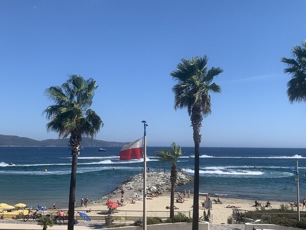 A beach with palm trees, a flag, and people enjoying the seaside. - Magnificent air-conditioned apartment, sea view, elevator, Internet, Parking (Cavalaire-Sur-Mer)