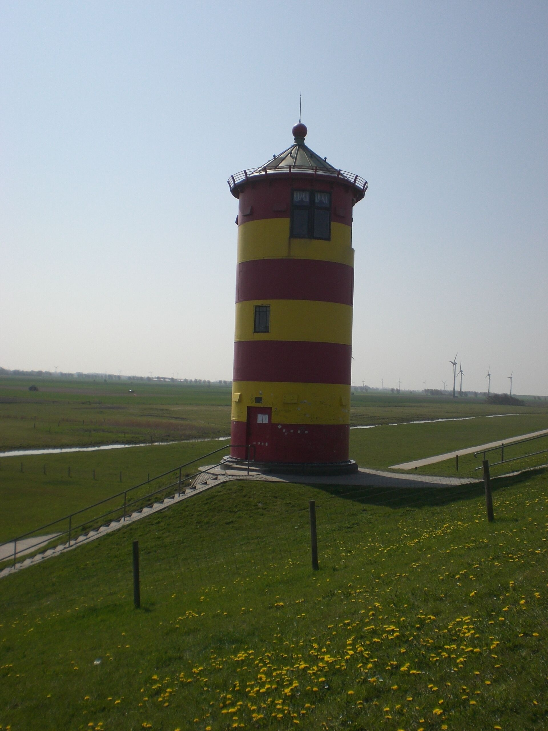 A cylindrical lighthouse with red and yellow stripes, situated on a grassy hill with a clear sky.