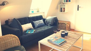 Ein gemütliches Wohnzimmer mit einem dunkelblauen Sofa, einem Korbstuhl, einem Couchtisch mit Büchern und einem Bücherregal an der Wand.