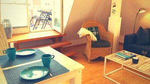 Ein gemütliches Wohnzimmer mit einem Esstisch für zwei Personen, einem Korbstuhl mit Kissen, einem dunklen Sofa und einem Couchtisch mit Büchern und einem Glas.