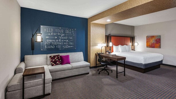 A hotel room with a gray sofa, a small wooden table, a desk, and a bed with white bedding. There is a motivational quote on the wall and a colorful abstract painting above the bed.