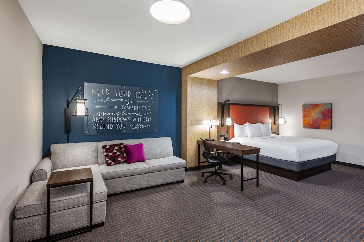 Hotelkamer met een grijze bank, een klein houten tafeltje, een bureau en een bed met wit beddengoed. Op de muur staat een motiverende quote en boven het bed hangt een kleurrijk abstract schilderij.