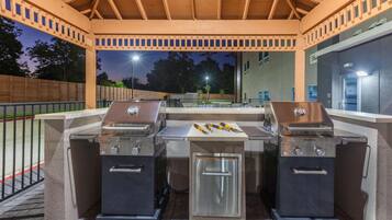 Area barbecue esterna con due griglie e un piccolo frigorifero, circondata da una pergola in legno e un muro in mattoni.