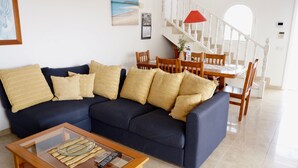 A living room with a dark blue sofa, multiple cushions, a glass coffee table with a remote control, a dining area with wooden chairs and a table, and a staircase with white railings. - Spacious house with 3 bedrooms, 2 bathrooms, swimming pool (Costa Calma)