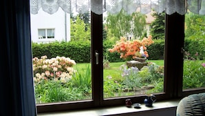 Ein Fenster mit Spitzenvorhängen, Blick auf einen Garten mit Blumen und einer Statue, und ein kleiner Tisch mit zwei Tassen.