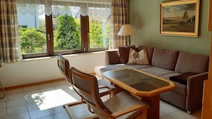 Ein Wohnzimmer mit Holzbalkendecke, einer Couch, einem Esstisch mit Stühlen, einem Fenster mit Vorhängen und einem Bild an der Wand.