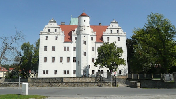 Ein großes, weißes Gebäude mit rotem Dach und zahlreichen Fenstern, umgeben von Bäumen und einer Steinmauer.
