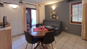 Ein Essbereich mit einem Holztisch, vier Stühlen, einer roten Tischdecke und einer Weinflasche. Im Hintergrund befindet sich ein graues Sofa und ein Fenster mit Jalousien.