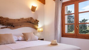 A bedroom with a large wooden headboard, a bed with white linens, a hat on the bed, a nightstand with a lamp, and a window with a view of trees. - Villa with pool  Pollensa - Can Roig Guillem (Pollensa / Pollença)