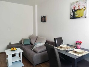 A modern living room with a grey sofa, a small white side table with fruit, a dining table with plates and cutlery, and a wall-mounted picture of flowers.