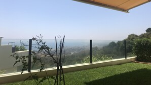 A balcony with a clear view of the cityscape, a grassy area, and a glass railing.