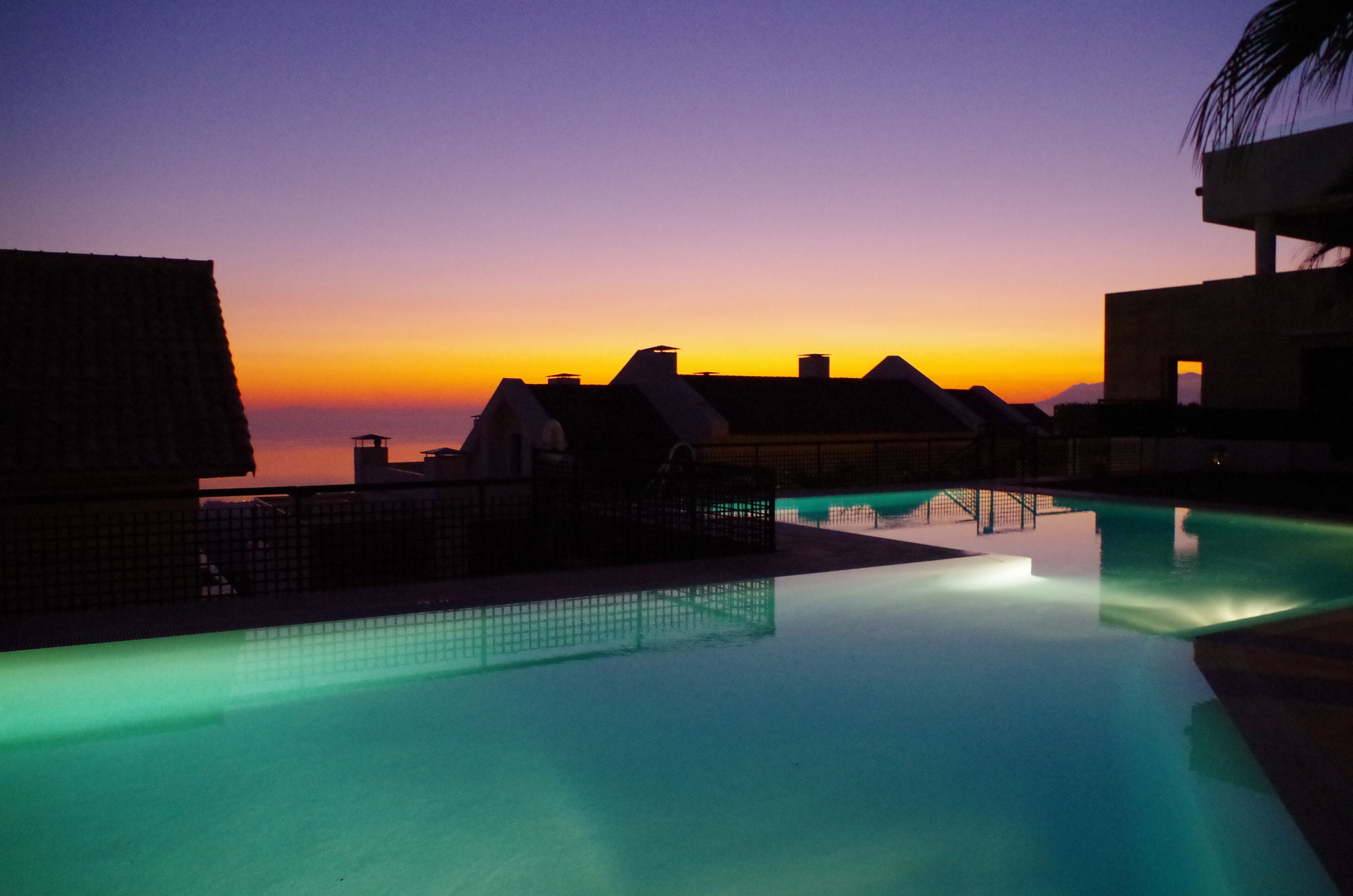 Área de piscina com vista para o pôr do sol, telhados salientes e superfície de água calma.