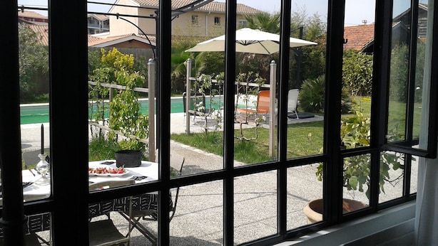 Vista a través de una ventana que muestra una mesa y sillas afuera, una piscina y un edificio al fondo.