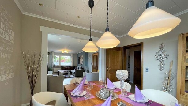 Uma sala de jantar com uma mesa de madeira posta para refeição, com guardanapos roxos, cadeiras brancas e um candelabro.