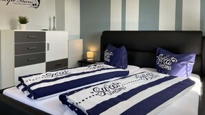 Quarto de hotel com uma cama com roupa de cama listrada, uma mesa de cabeceira com abajur, uma cômoda e uma parede com listras verticais e um letreiro decorativo.