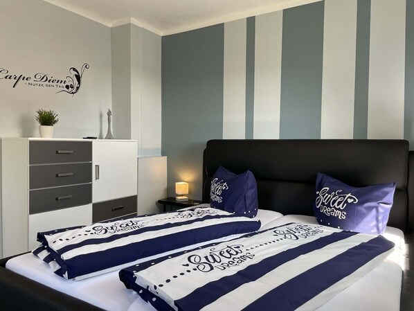 Habitación de hotel con una cama con ropa de cama a rayas, una mesita de noche con lámpara, un aparador y una pared con rayas verticales y un letrero decorativo.