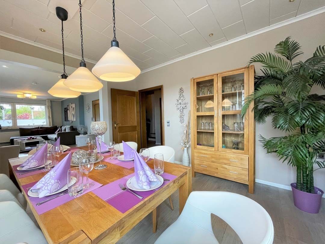 Un comedor con una mesa de madera dispuesta para cuatro personas, rodeada de sillas blancas. Hay lámparas colgantes sobre la mesa.